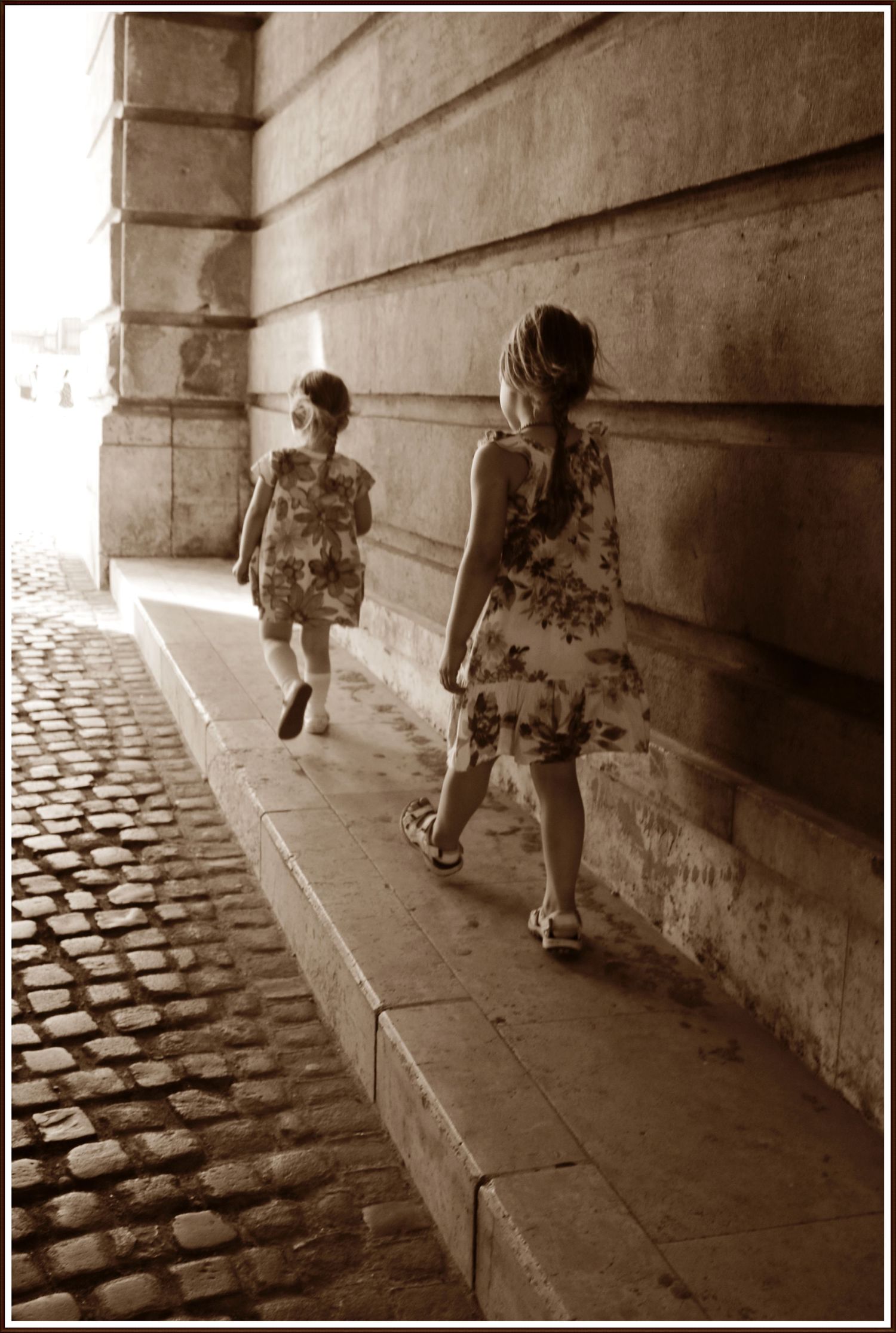 Two people in summer dresses walk along a cobblestone path beside a historic stone wall.
