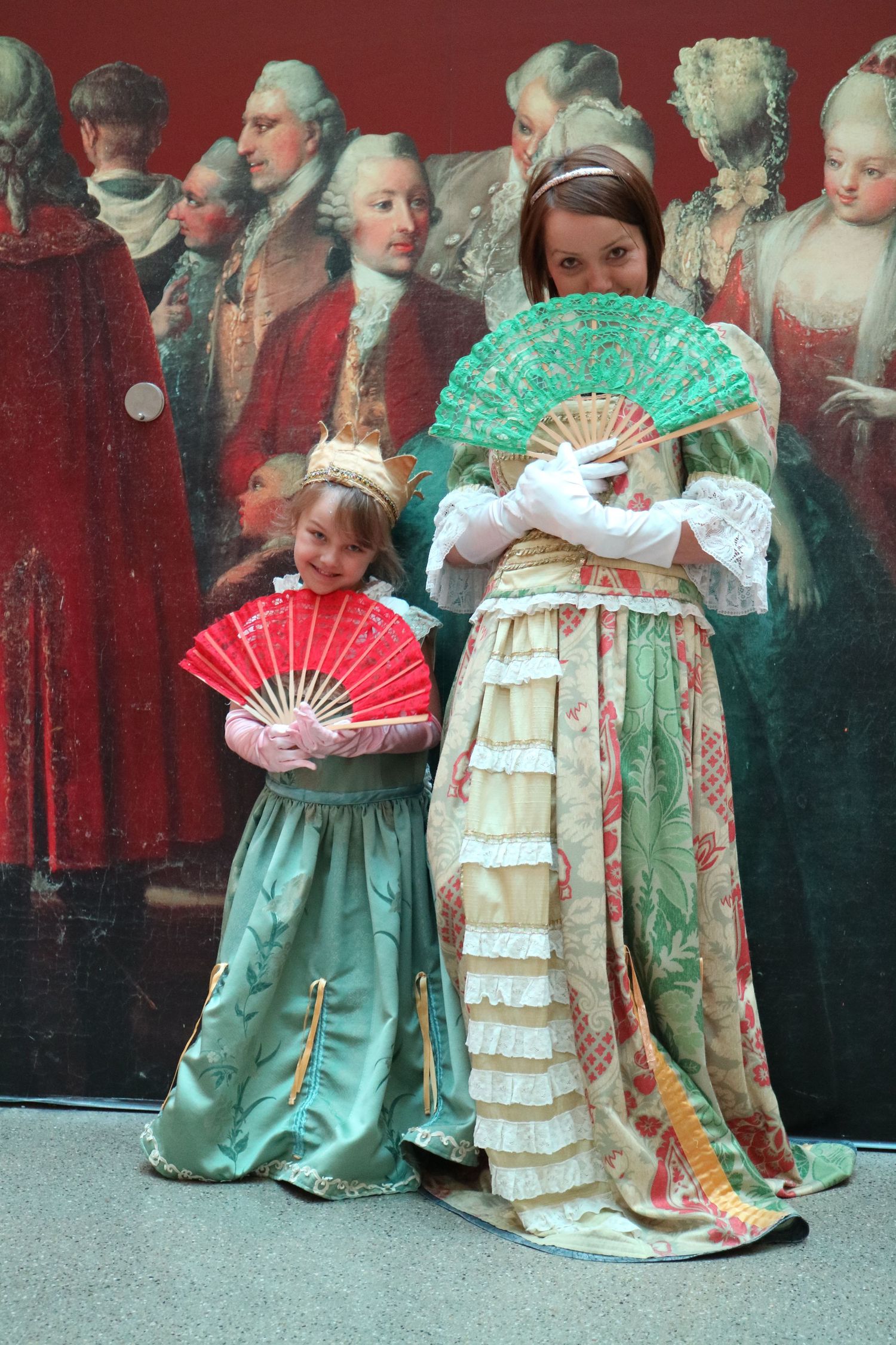 People in period costumes holding decorative fans pose against a backdrop of historical portraits.