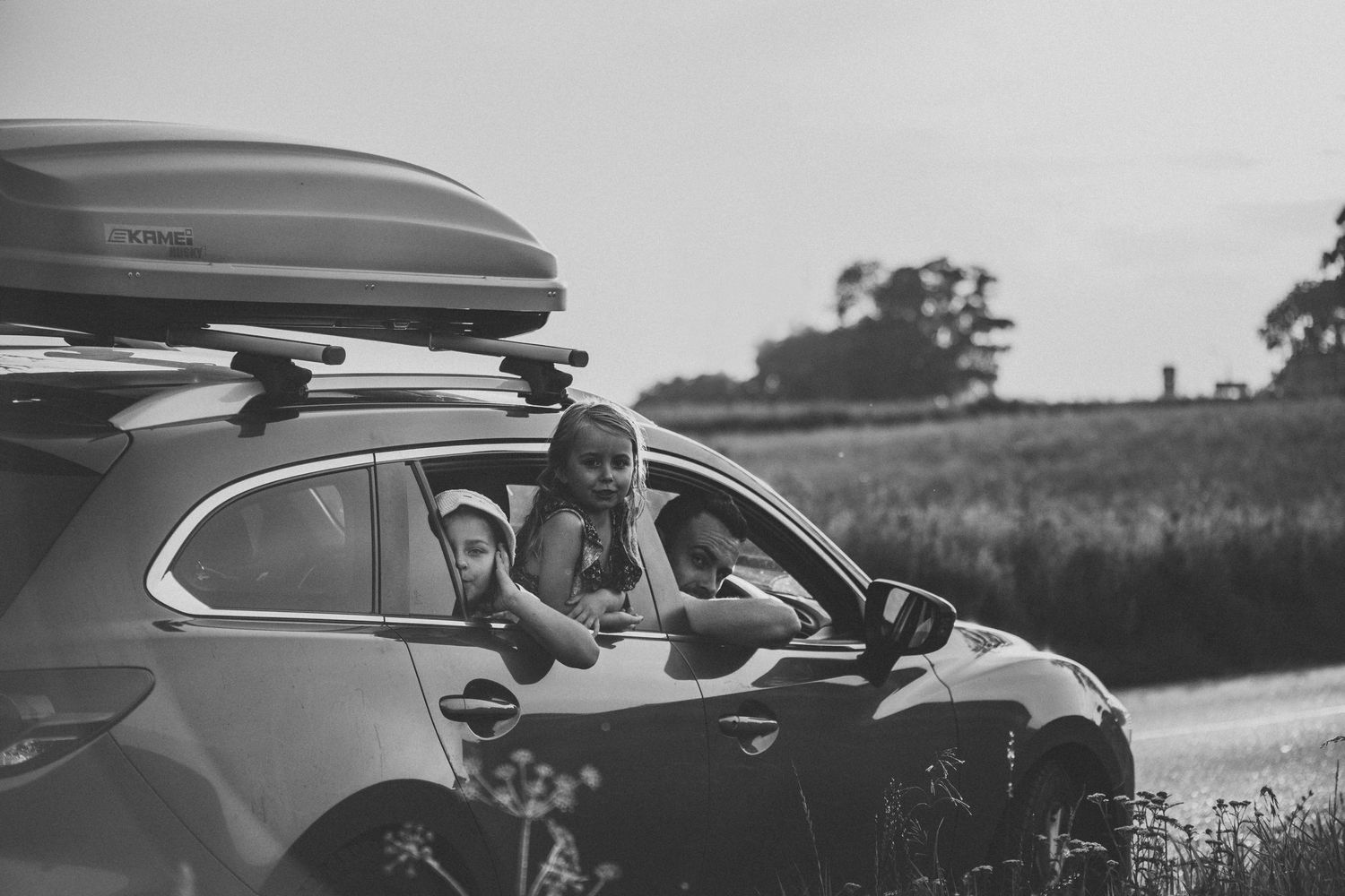 Black and white image of passengers in a car with roof cargo box during a road trip.