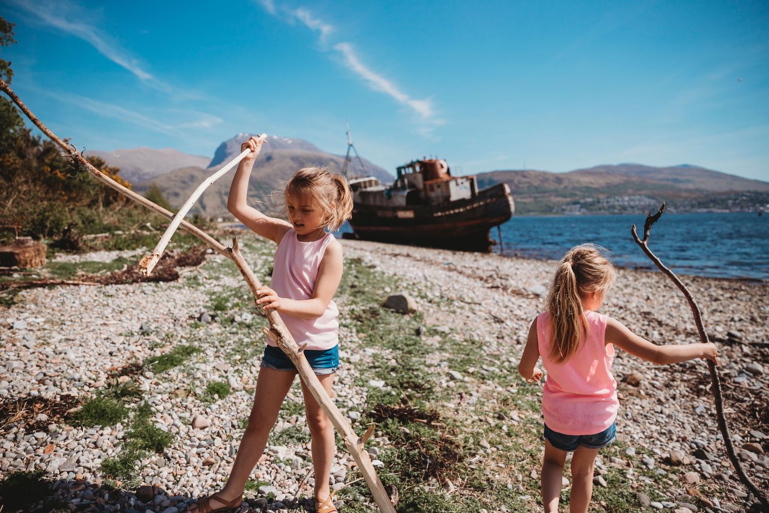 Two children play on a rocky beach with an abandoned boat in the background.