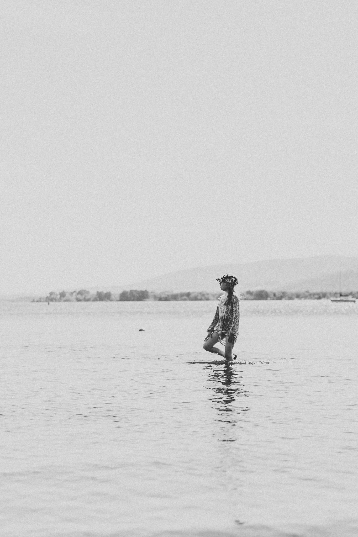 A solitary figure stands in shallow water wearing a dress and hat against a minimalist landscape.