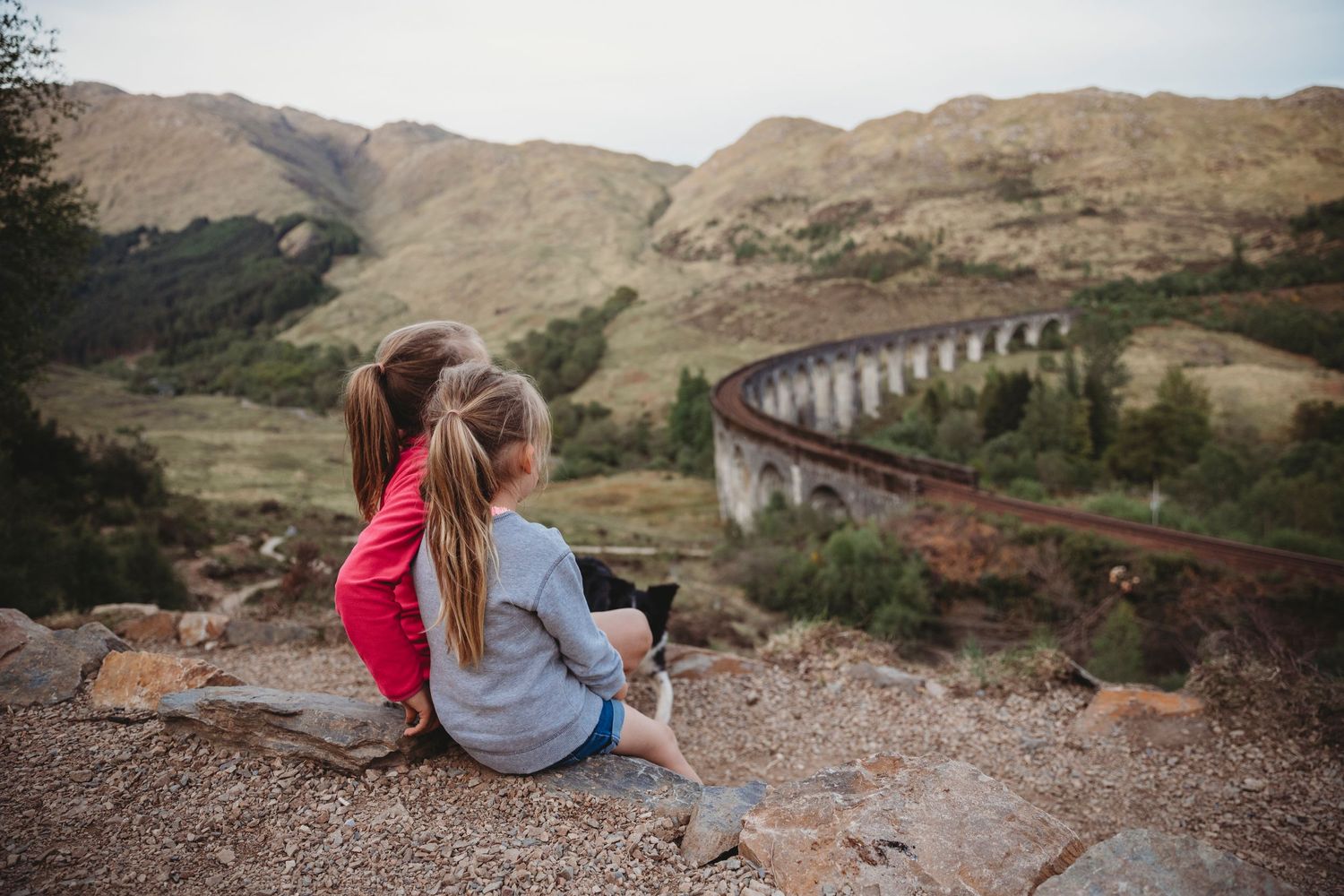 Two seated figures overlook a scenic railway viaduct winding through mountain landscape at sunset.