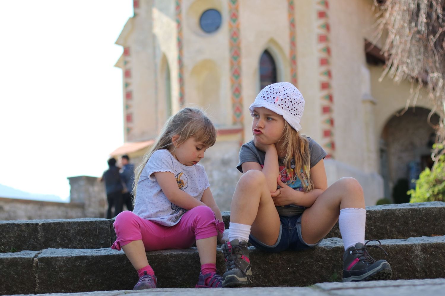 Two people sit and chat on stone steps near a decorative church facade.