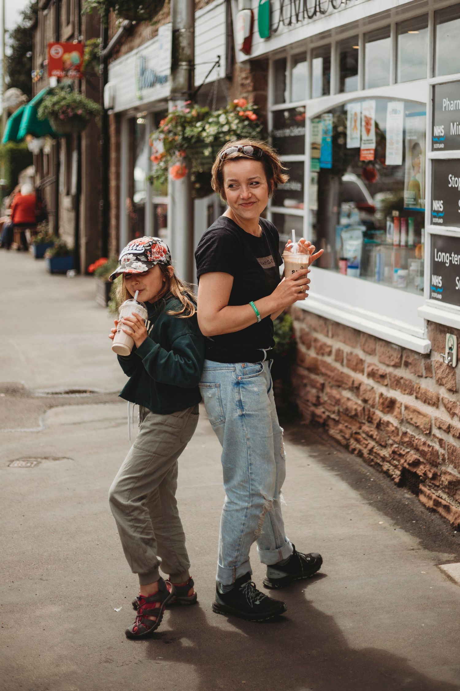 Two people enjoying drinks while standing on a sidewalk lined with flower baskets and storefronts.