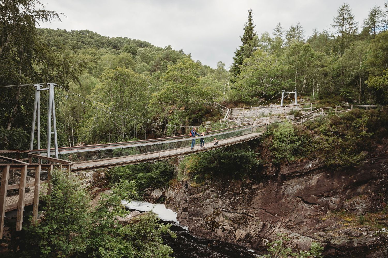 A wooden suspension bridge spans across a rocky gorge in a lush forest setting.