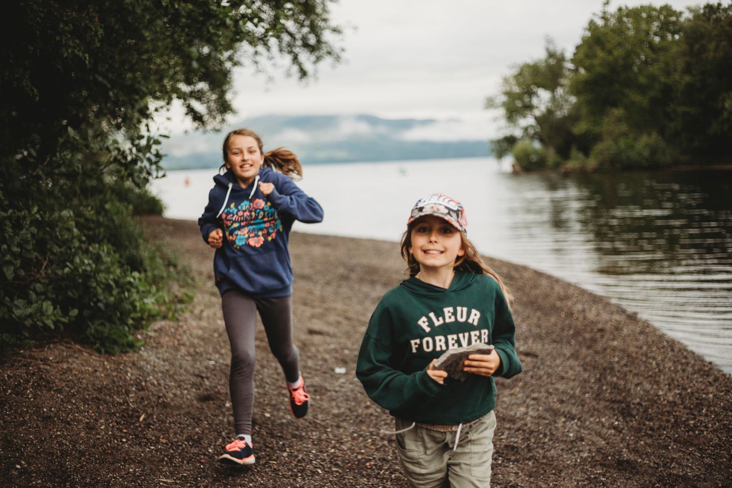 Children in cozy sweatshirts playing on a lakeside trail surrounded by natural scenery.