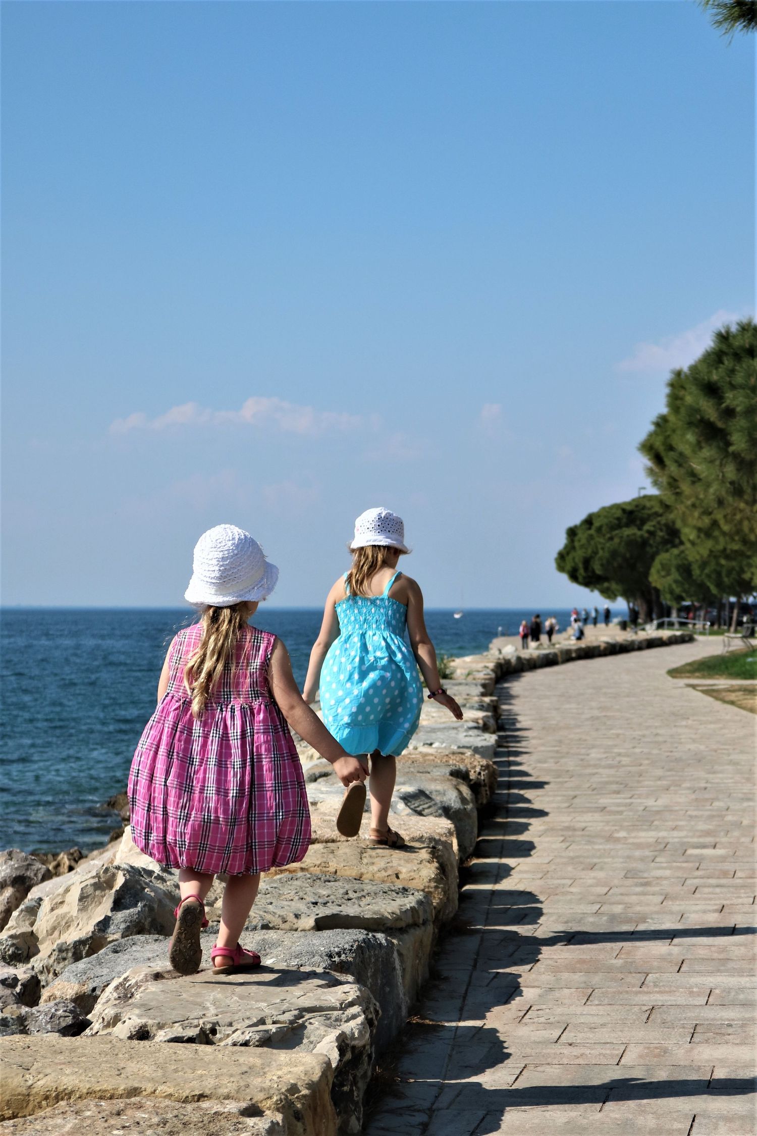 Young girls in summer dresses explore a seaside walkway lined with rocks and trees.