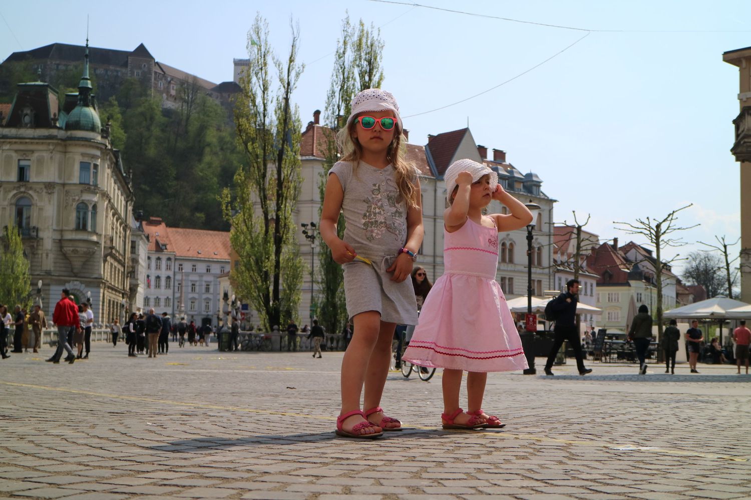 Children in summer outfits stand in a European town square with historic buildings.