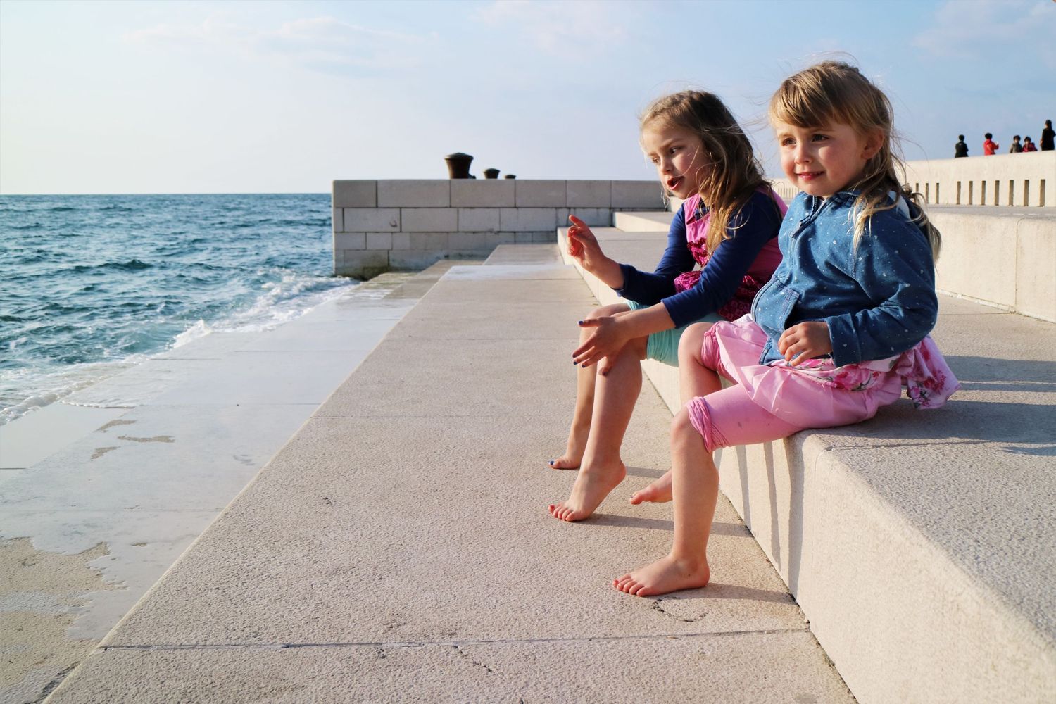 Two children sit on a concrete seawall by the ocean, dangling their bare feet over the edge.
