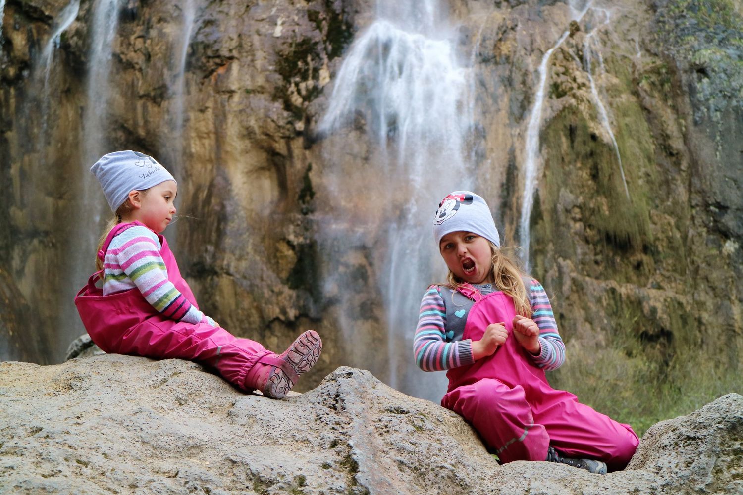 Two people in pink outfits sitting on rocks near a cascading waterfall.