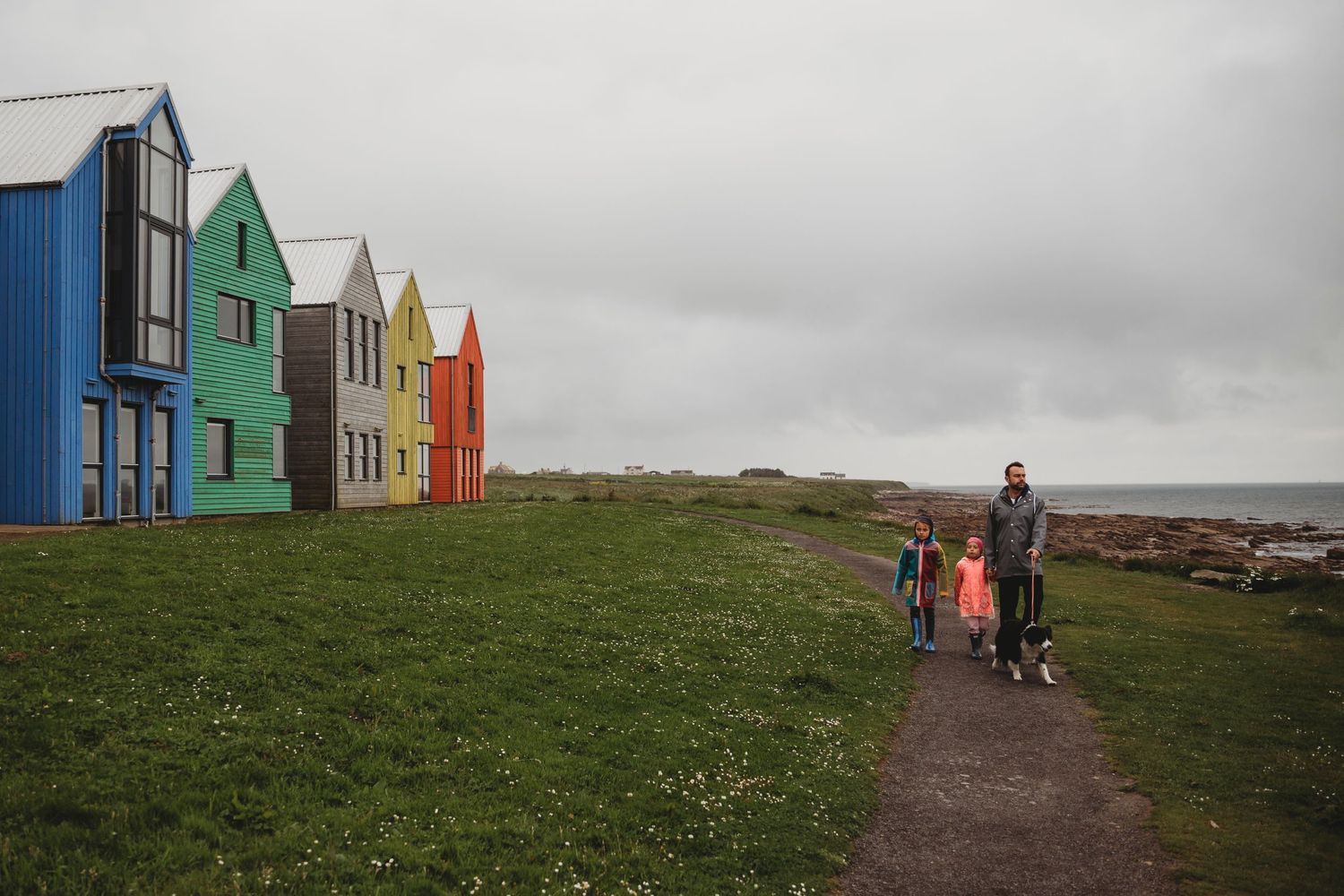 Colorful beach houses line a coastal path under cloudy skies.