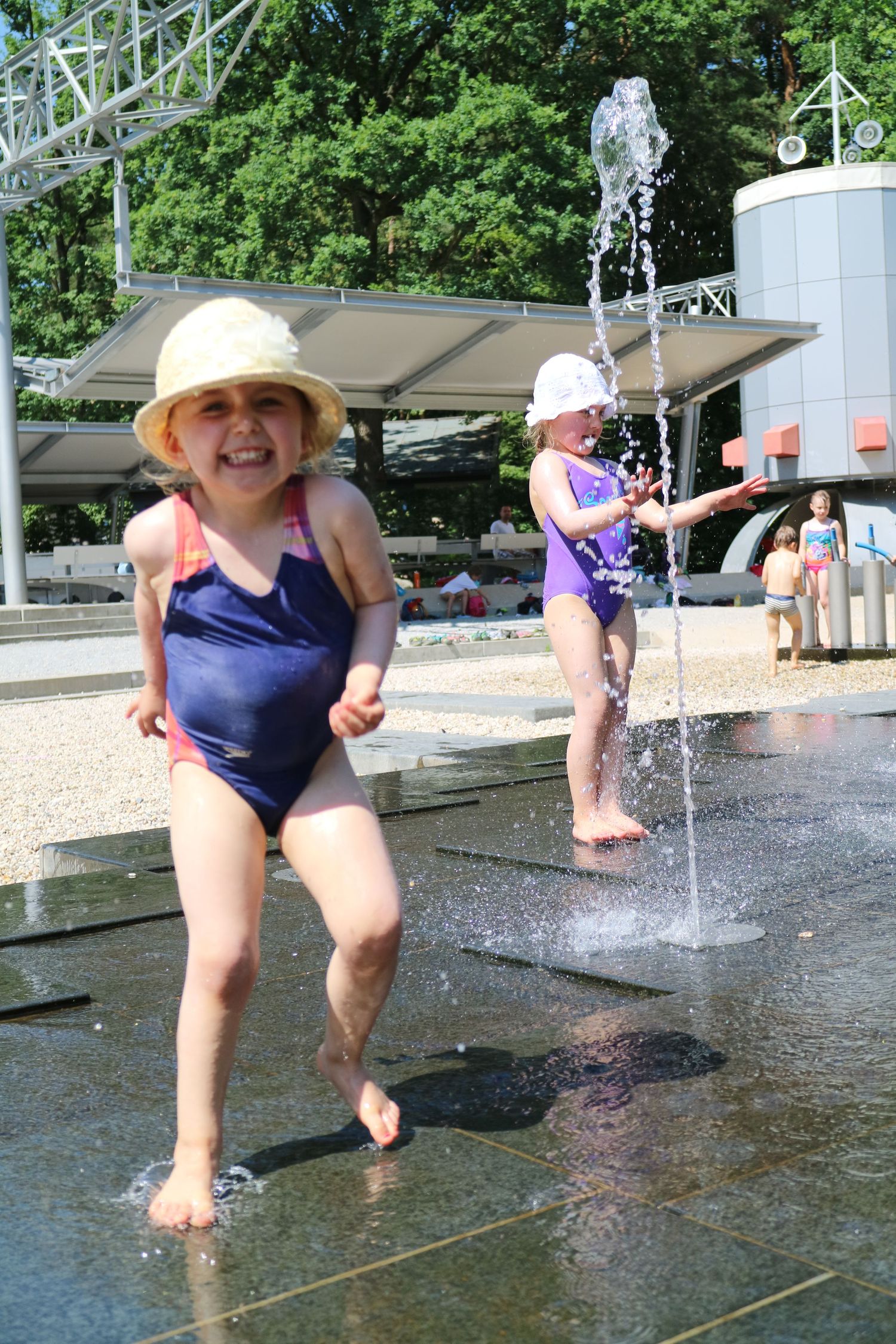 Children playing in an outdoor water fountain splash pad on a sunny summer day.
