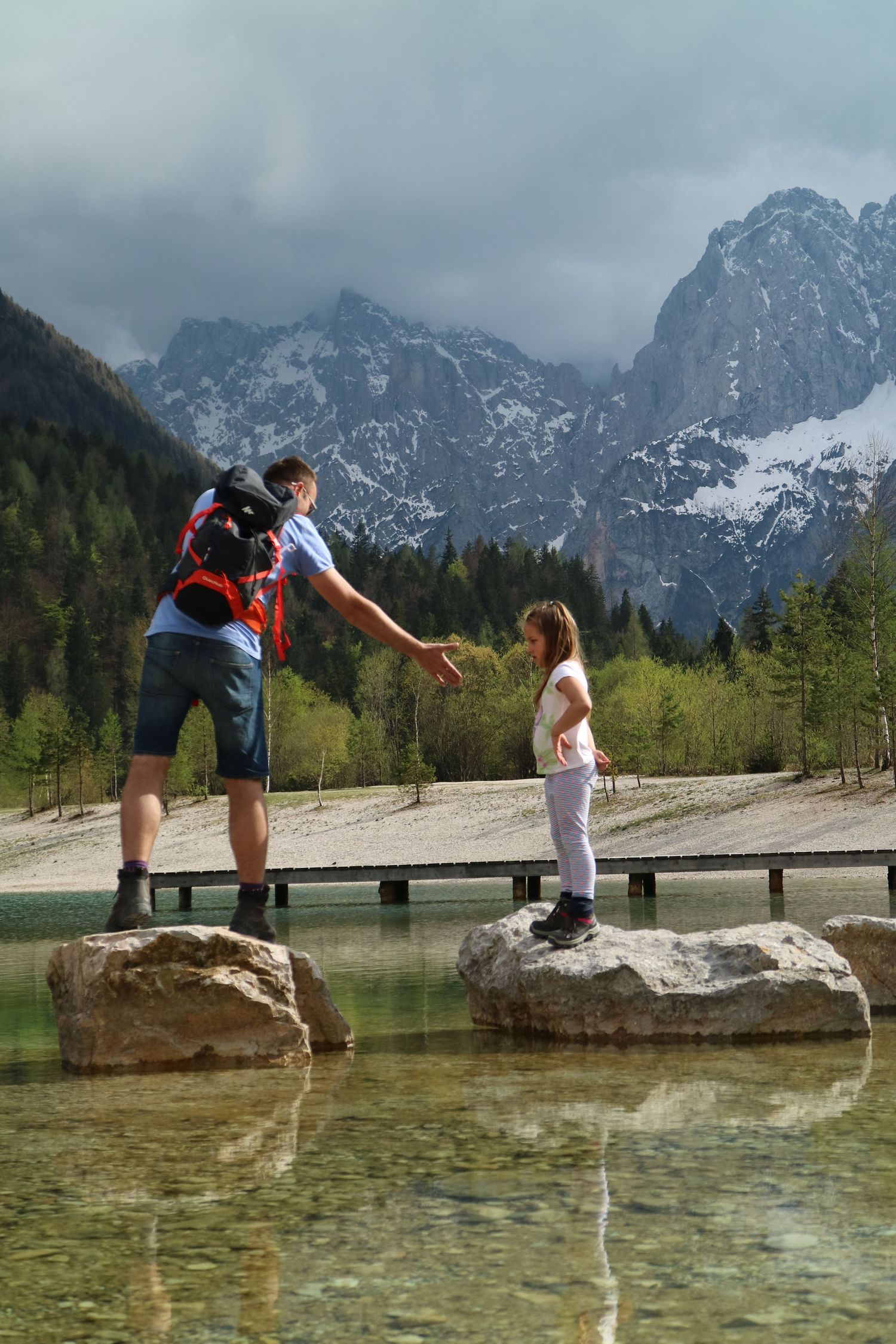 Hikers balance on rocks over a mountain lake with snow-capped peaks in the background.