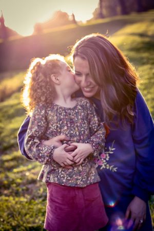 a daughter gives her mother a kiss on the cheek while sunlight streams behind them