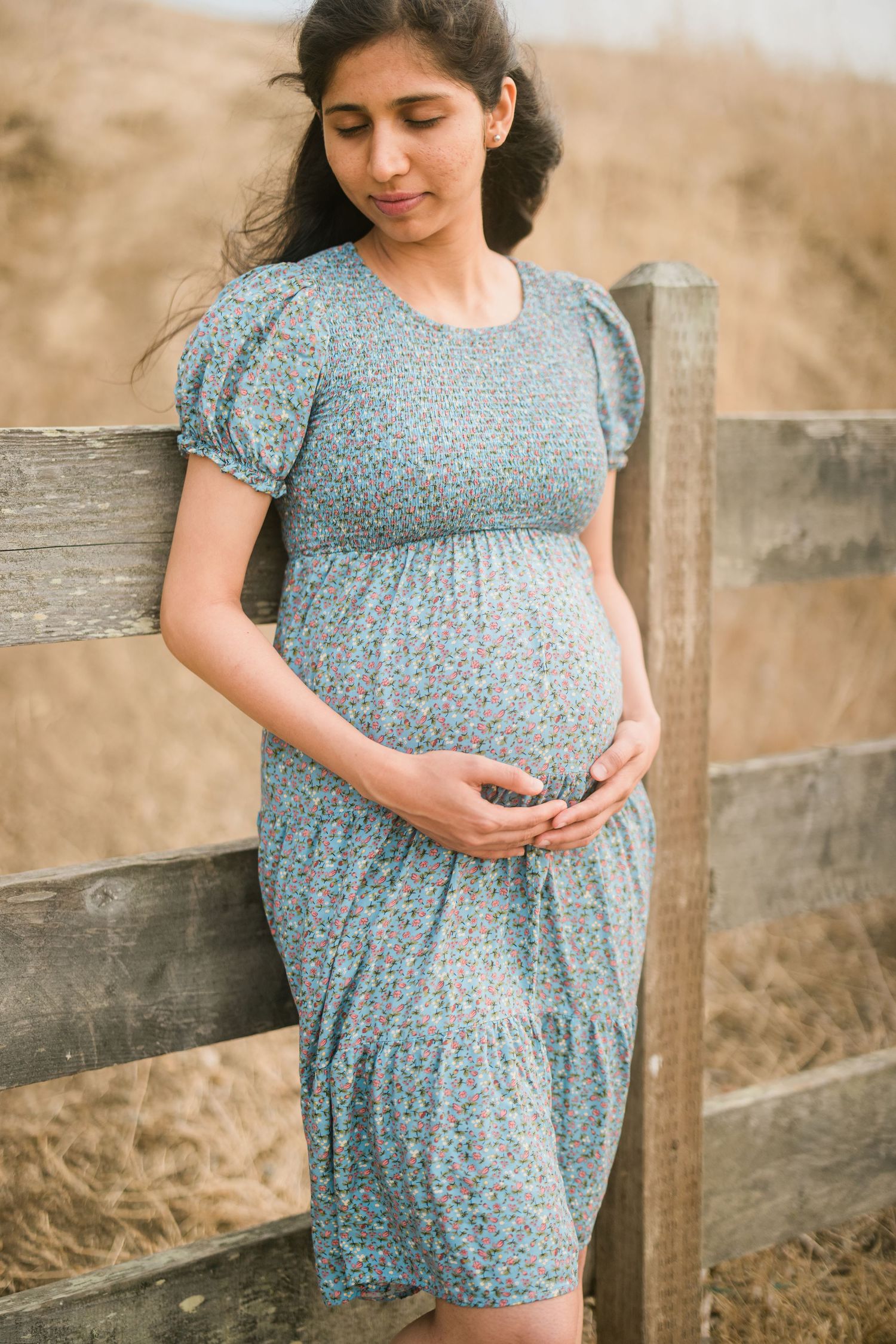 Maternity portrait of woman in blue floral dress leaning against wooden fence.