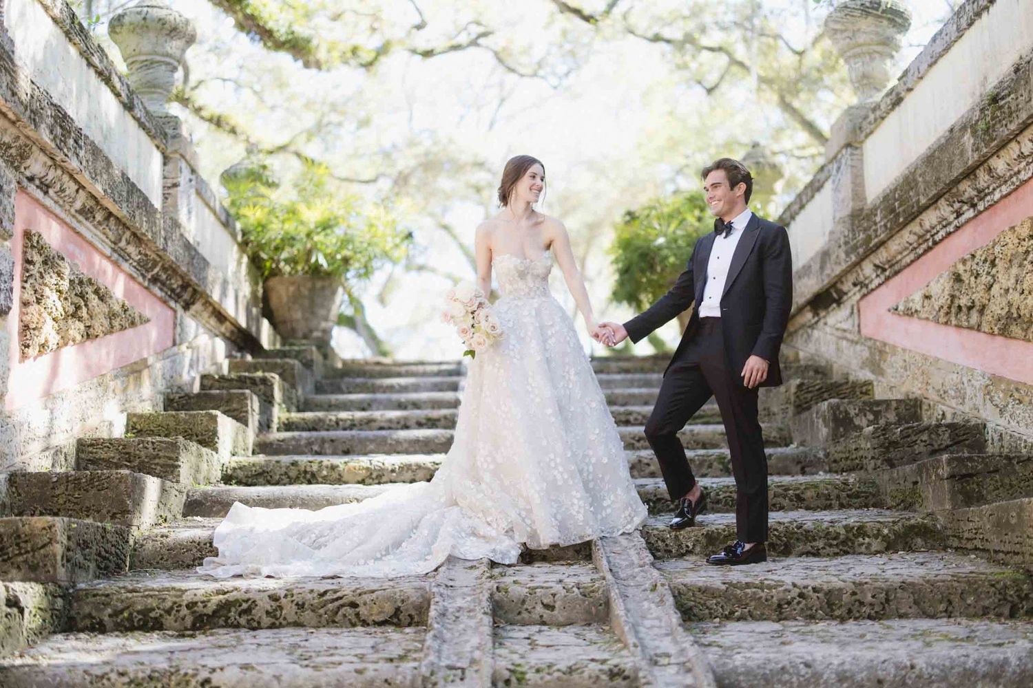 A couple in wedding attire poses on grand stone steps amongst overhanging trees and ornate railings.