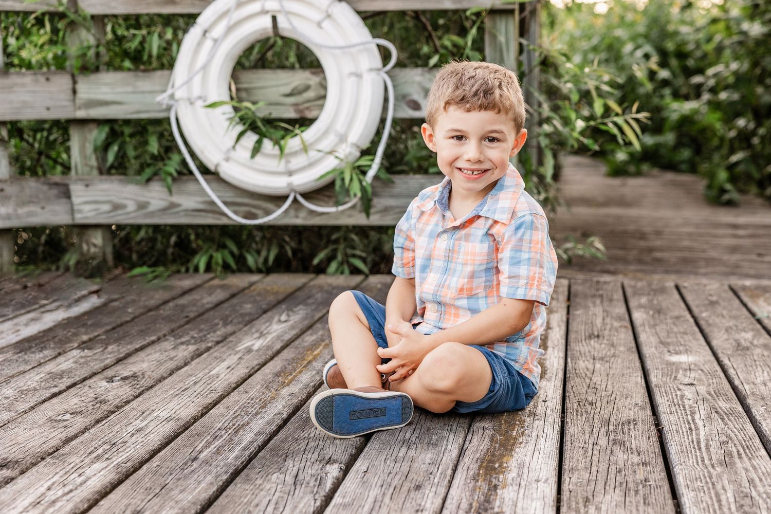 Young child sitting cross-legged on wooden deck with white life preserver decoration on wall.