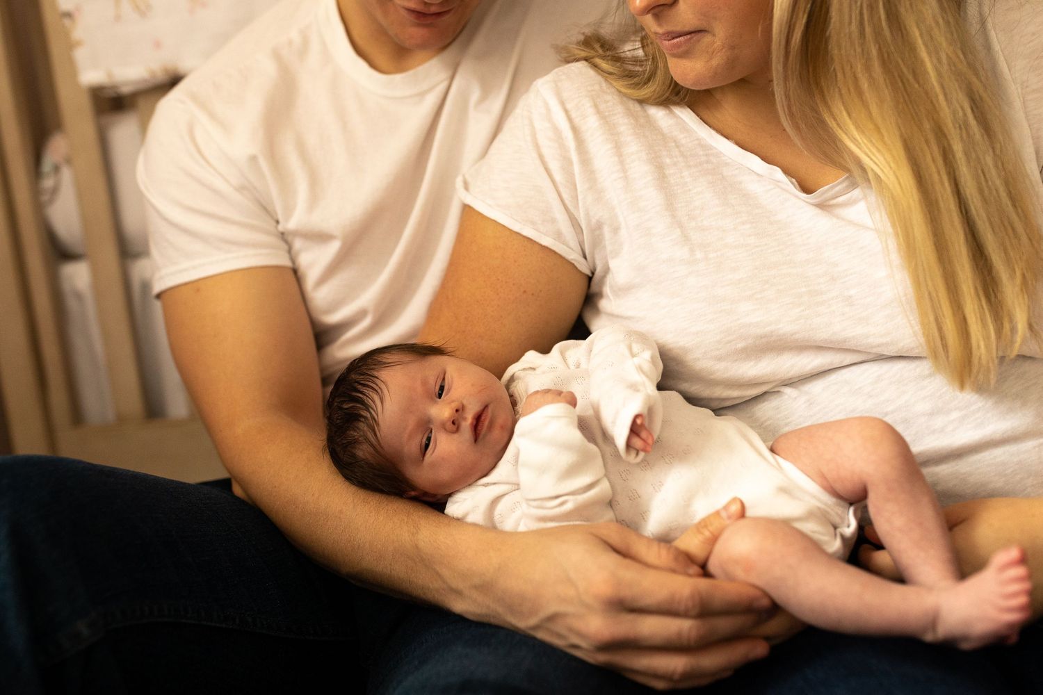 A baby in a white onesie rests peacefully in their parents' arms on a cozy couch.