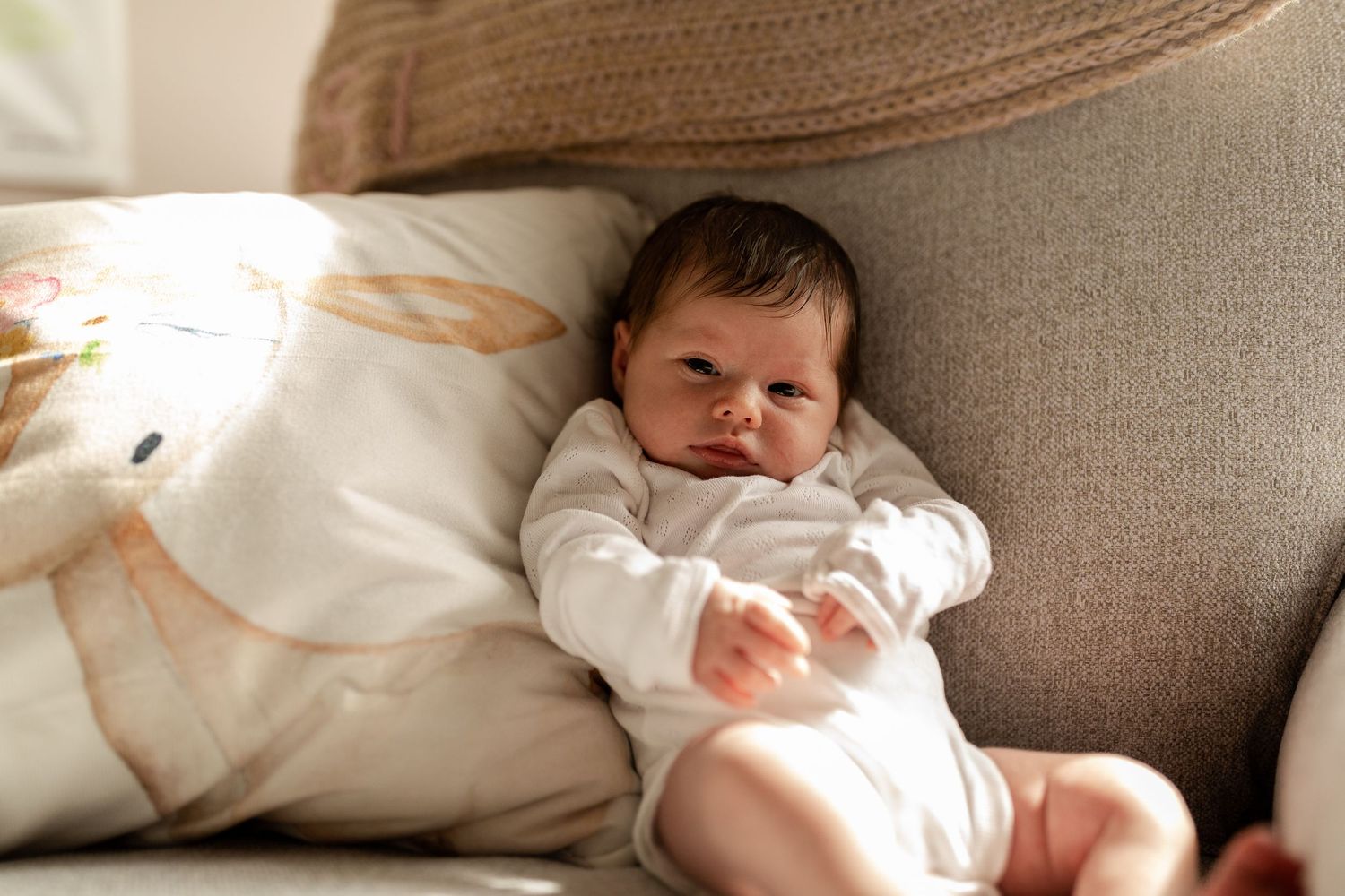 A baby in white clothing relaxes on a gray couch cushion.