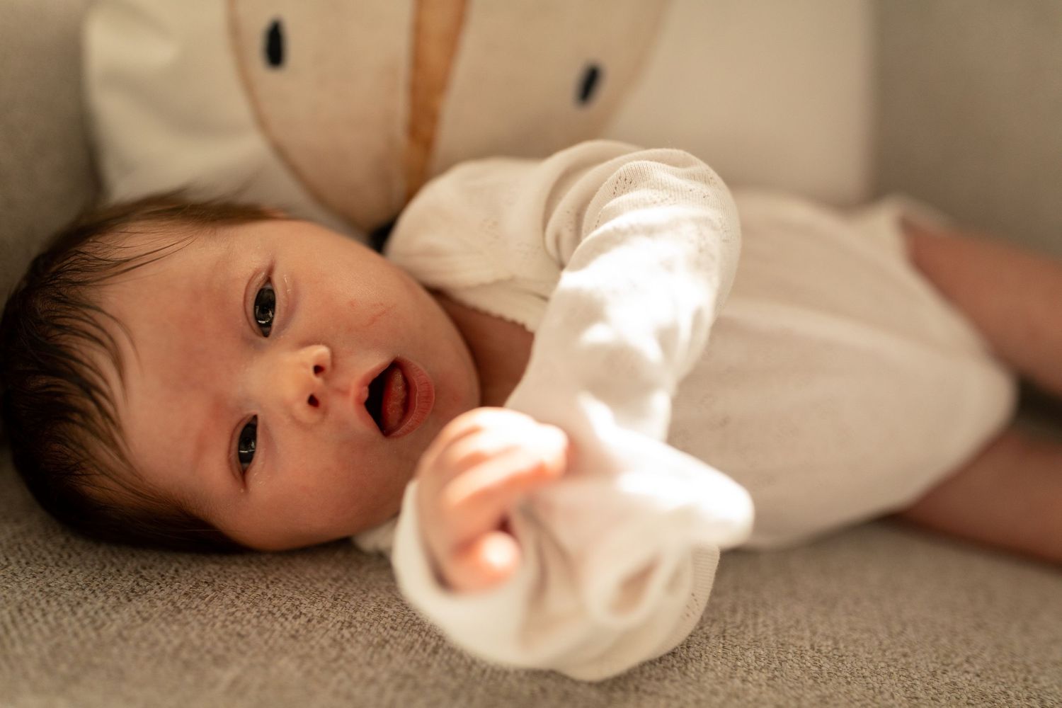 Baby wearing white clothing lies on beige couch with soft lighting.