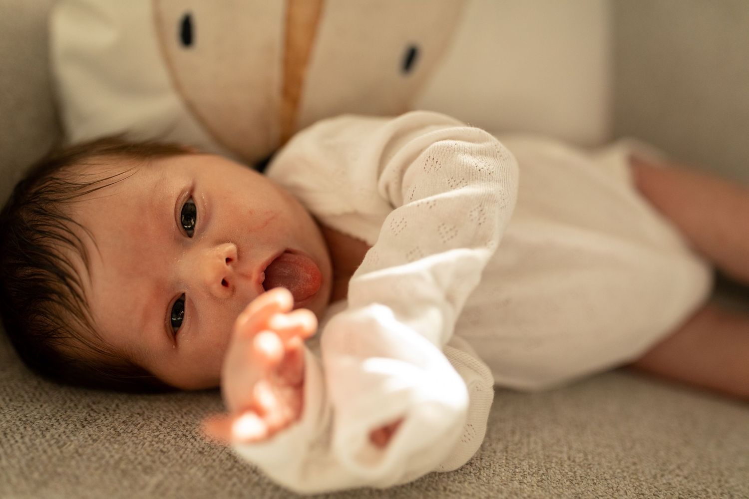 Baby in white clothing rests on couch with sunlight streaming across their face.