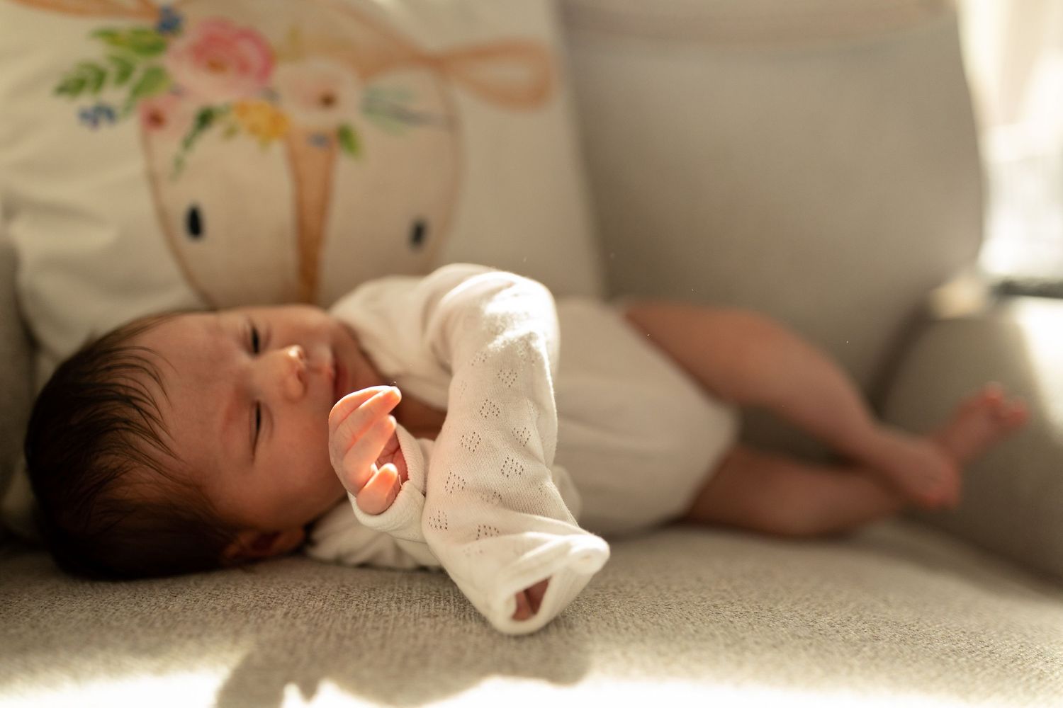 Baby in white outfit lies on couch with decorative pillow in background.