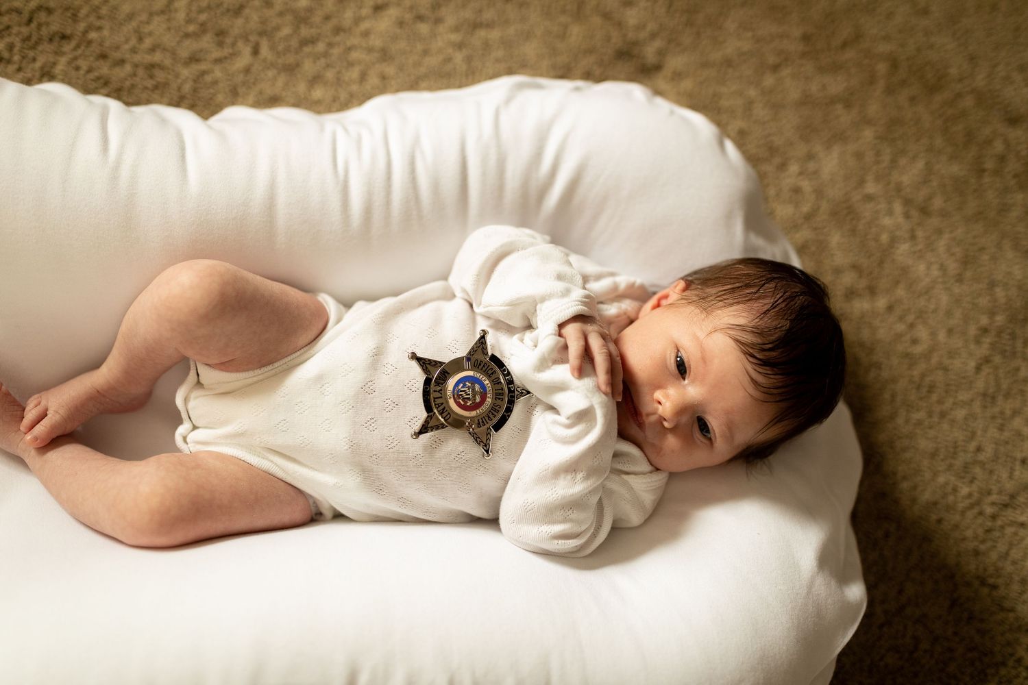 Baby wearing white onesie with sheriff badge lies on white cushion.