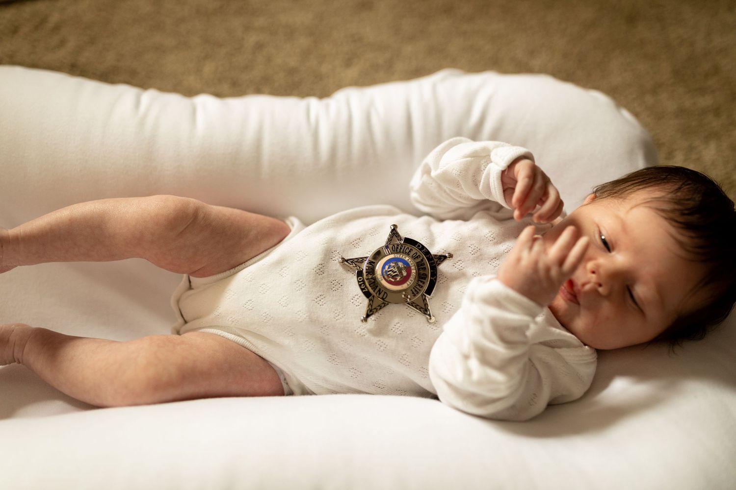 Baby wearing white onesie with sheriff badge stretches on white cushion.