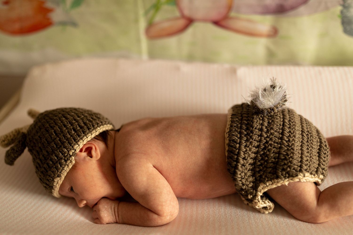 Baby wearing brown knitted hat and diaper cover on white blanket.