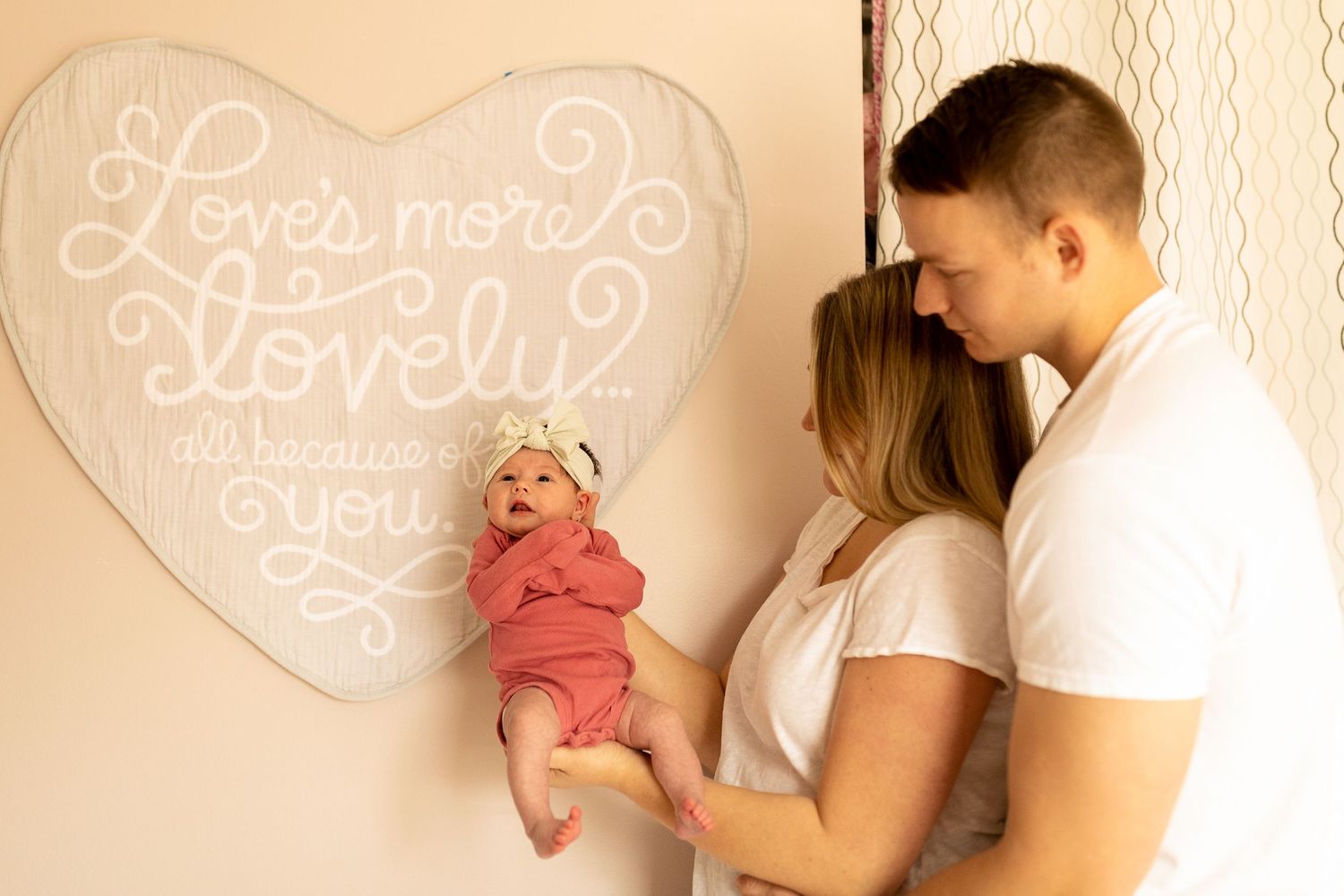Parents holding newborn baby against wall with decorative heart art.