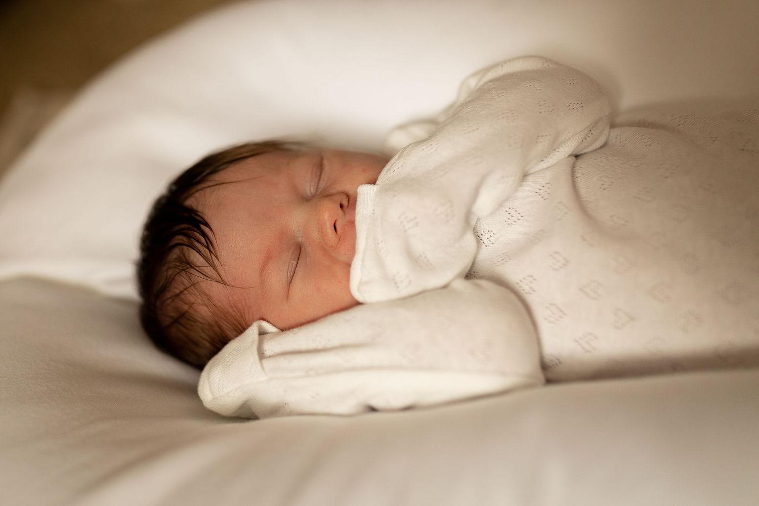 A sleeping newborn in white clothing against soft white pillows.