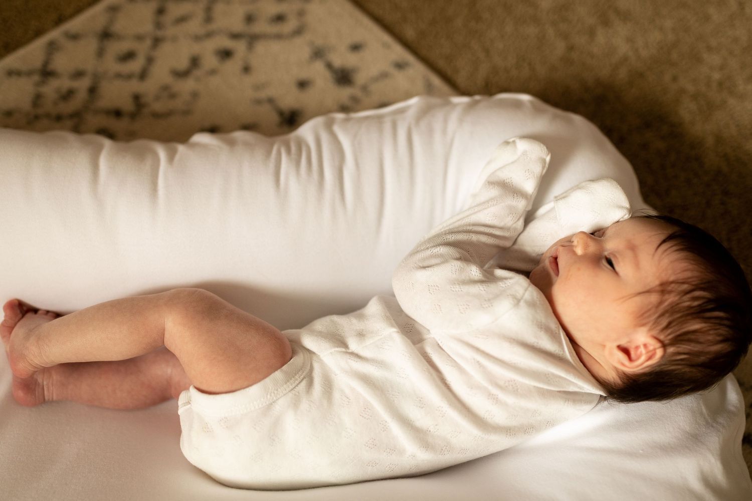 Baby wearing white clothing rests peacefully on beige bedding in warm lighting.