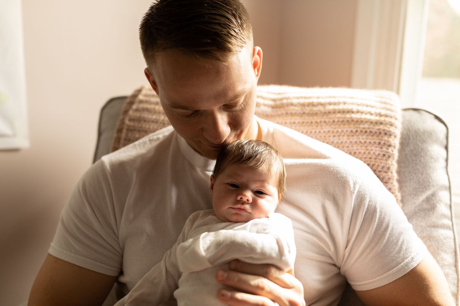 Parent in white t-shirt cradling newborn baby by sunlit window.