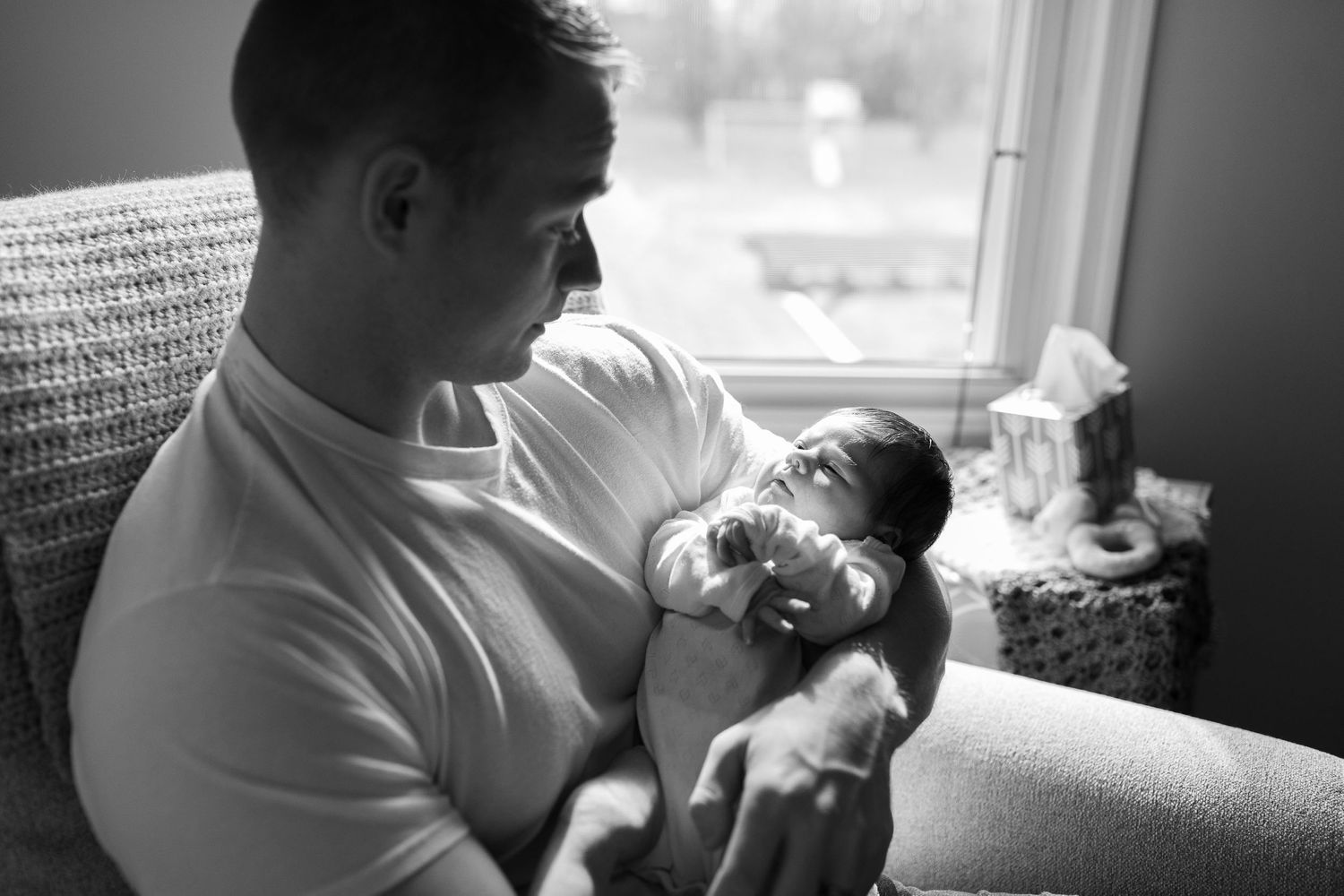 Black and white portrait of parent holding newborn baby near bright window.