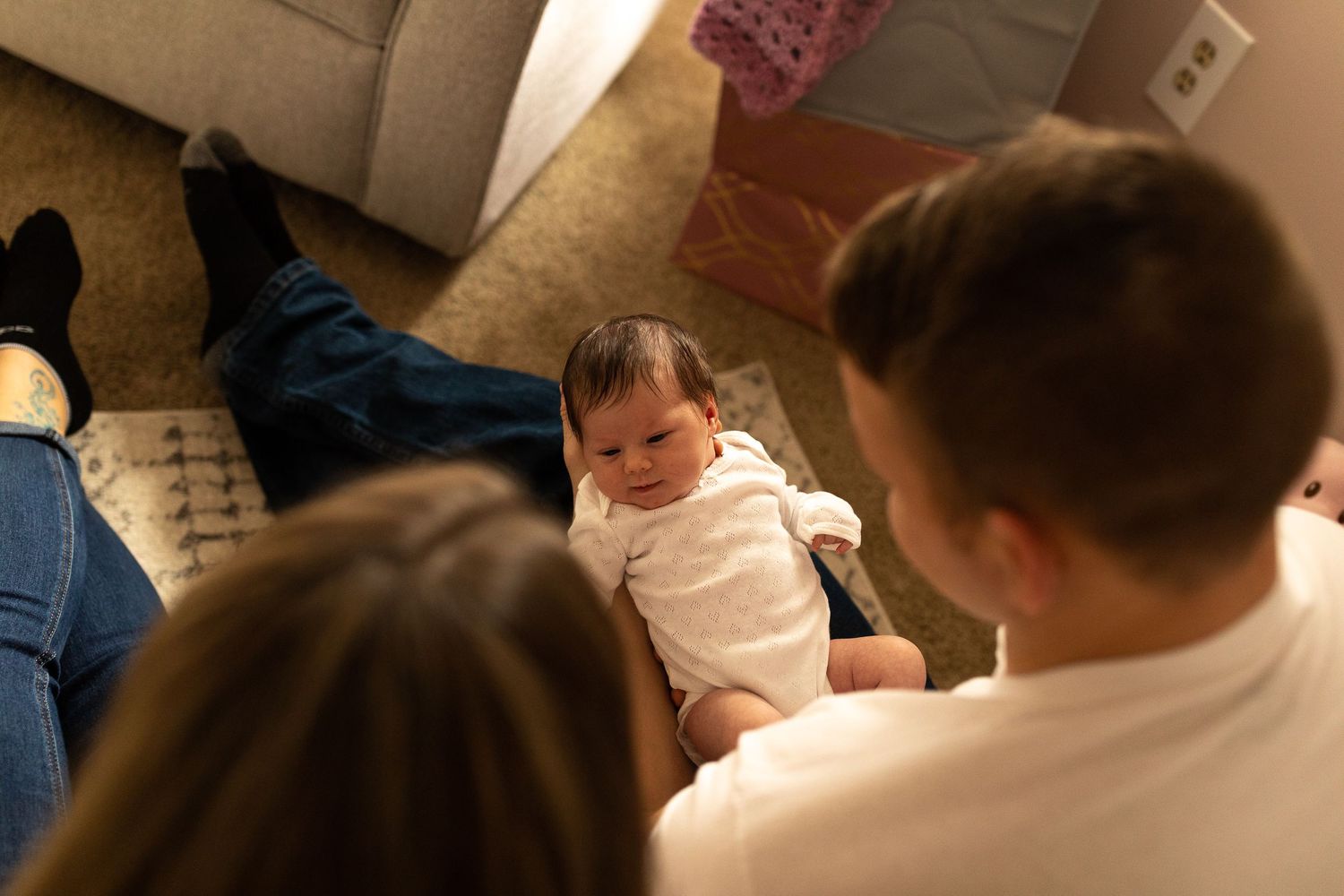 A baby in white clothing lays on dark fabric while being watched over.