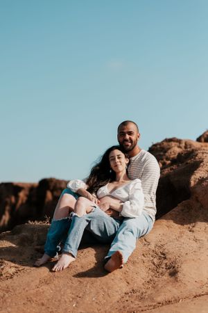 Couple lounging on rocky coastal cliff overlooking ocean on sunny day.