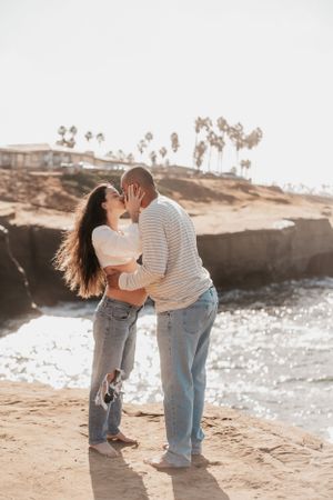Couple sharing playful moments on a beach with cliffs and palm trees.