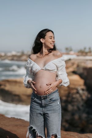 Maternity photos at sunset showing pregnant belly in white top and jeans against rocky coastal backdrop.