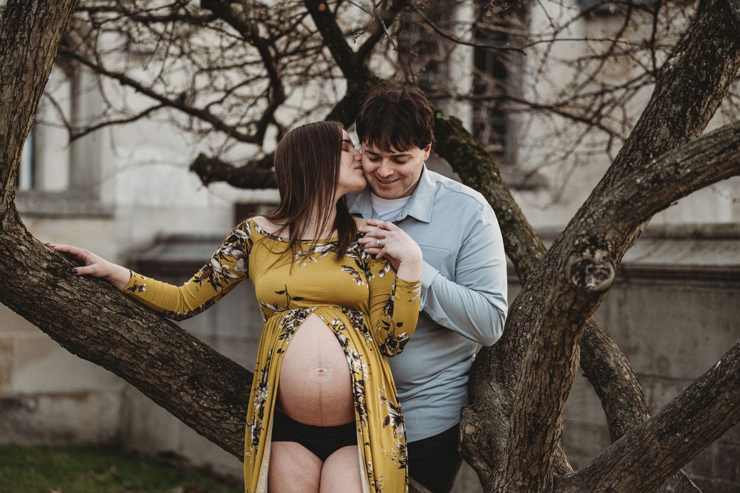 Pregnant couple embracing and kissing outdoors near bare tree branches.