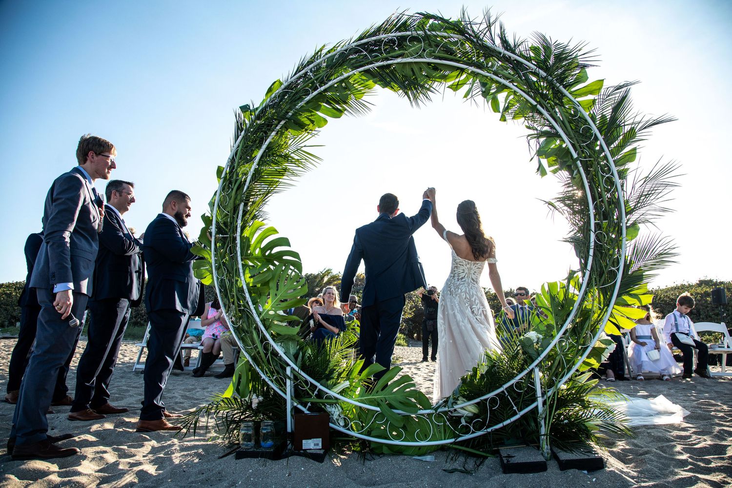 Wedding ceremony under circular greenery arch with silhouetted couple against sunset.