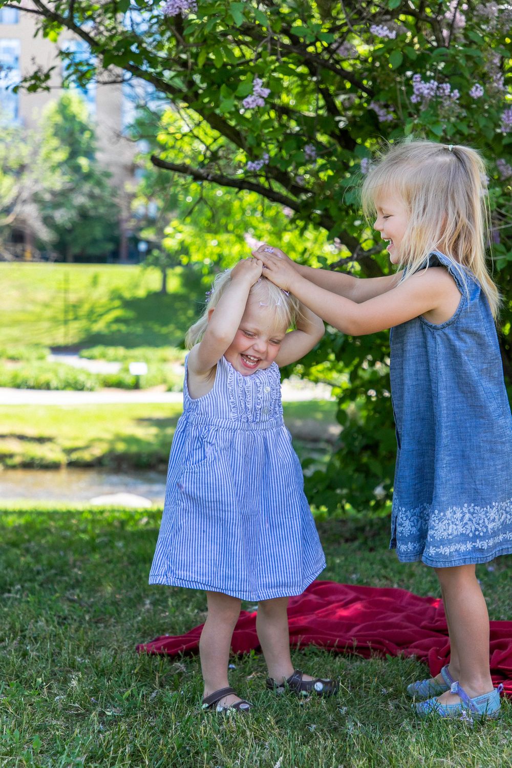 two young girls playing at a park by a tree with the older of the two placing petals on the younger girls head