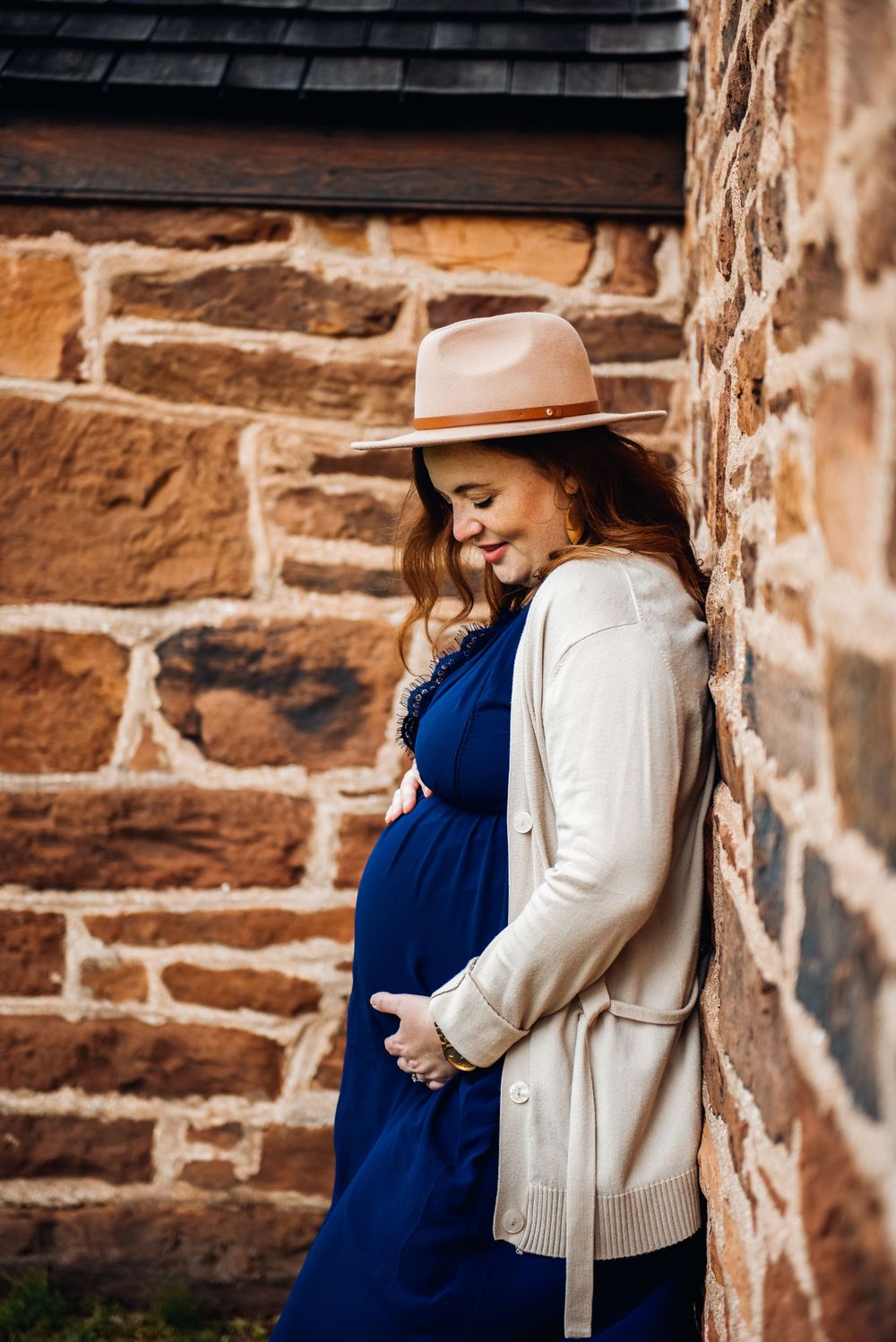 Pregnant woman in navy dress and hat stands against rustic stone wall.