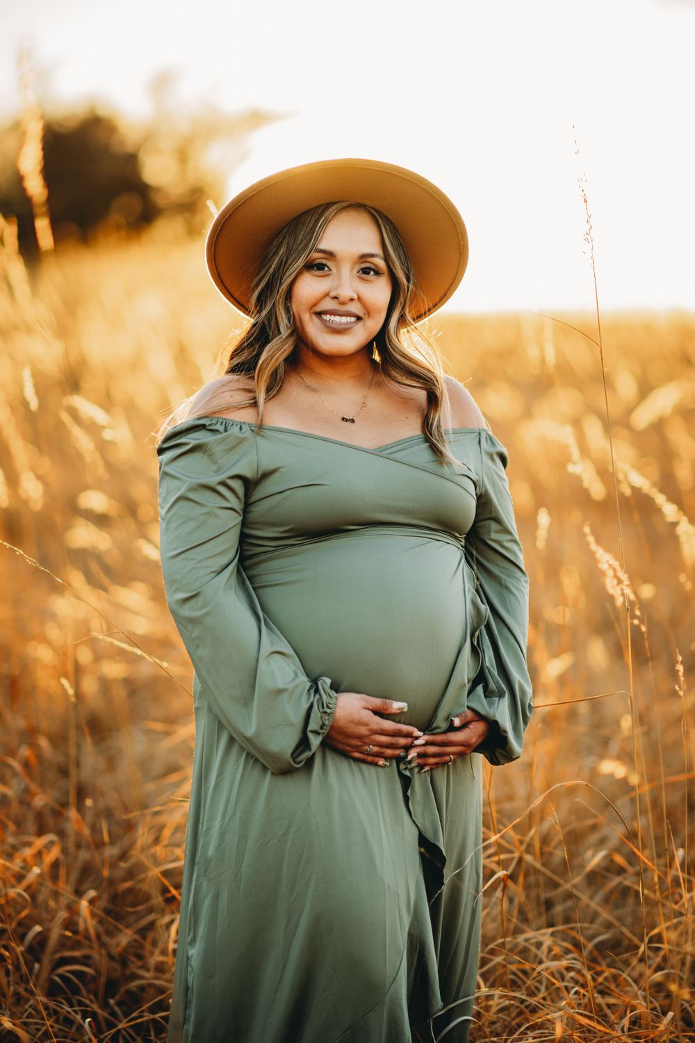 A maternity photo of a woman wearing a hat and green dress in a sunlit field.