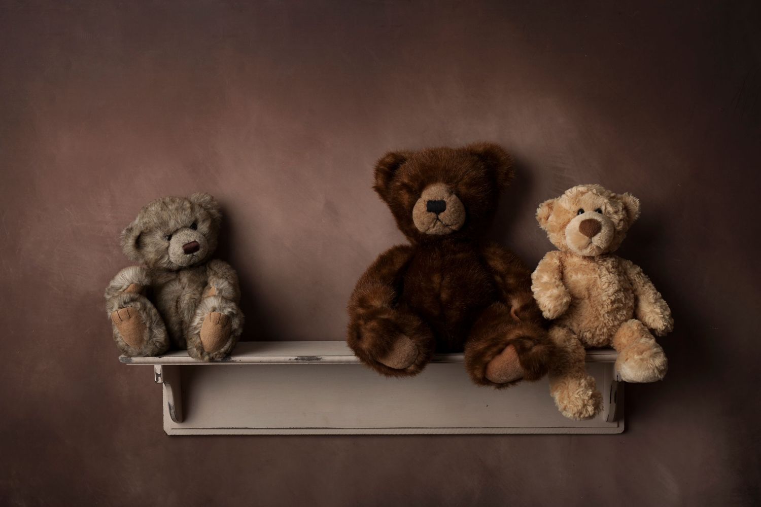 Three vintage teddy bears sitting on wooden shelf against brown background.