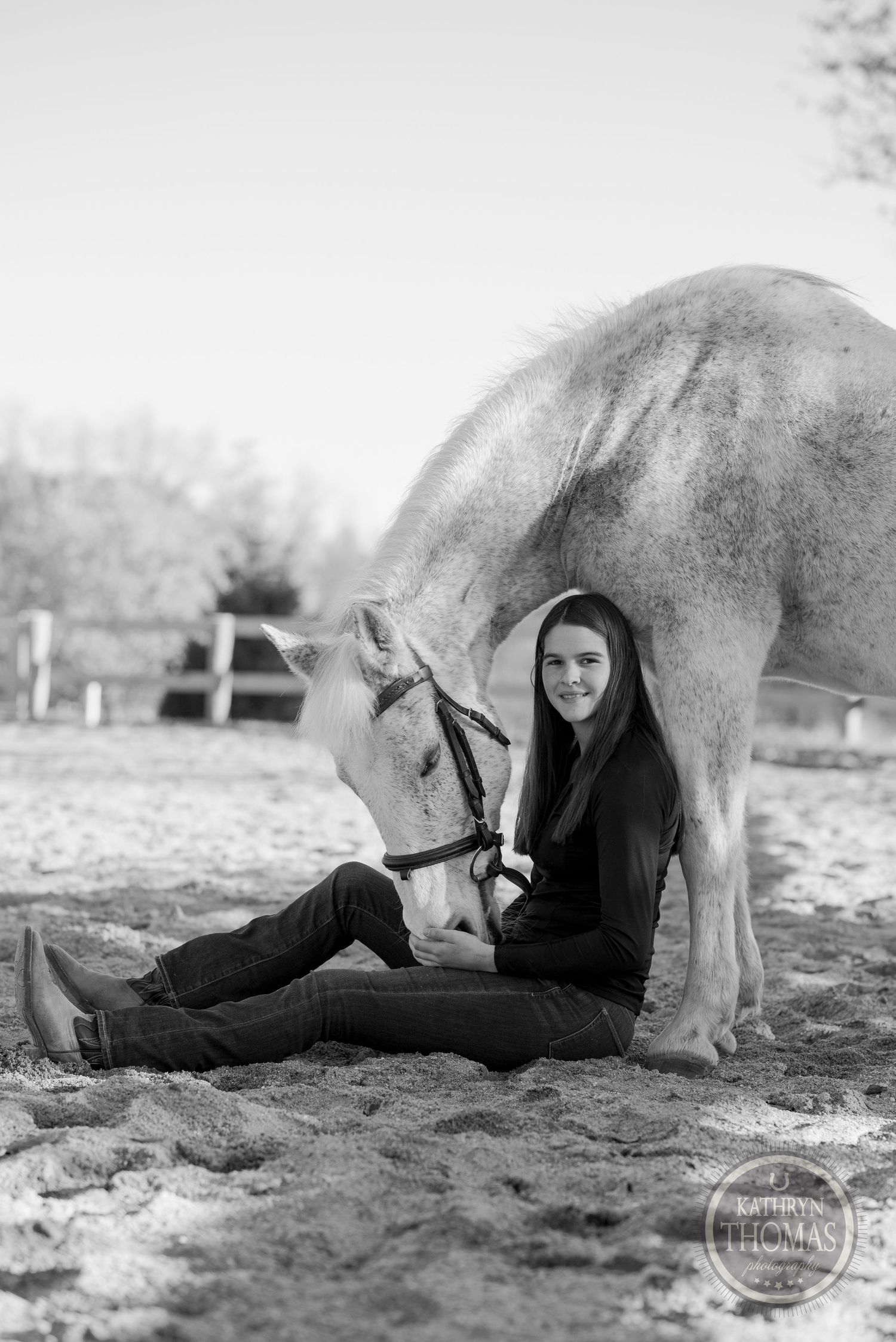 Black and white portrait of person relaxing next to a white horse in a sandy paddock.