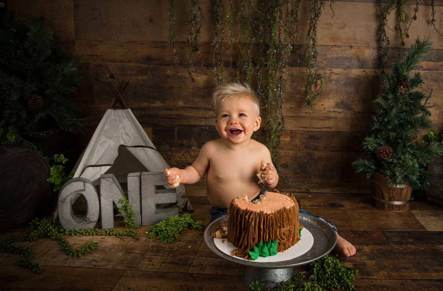 Joyful baby sits with a rustic woodland-themed smash cake and decorative ONE letters.