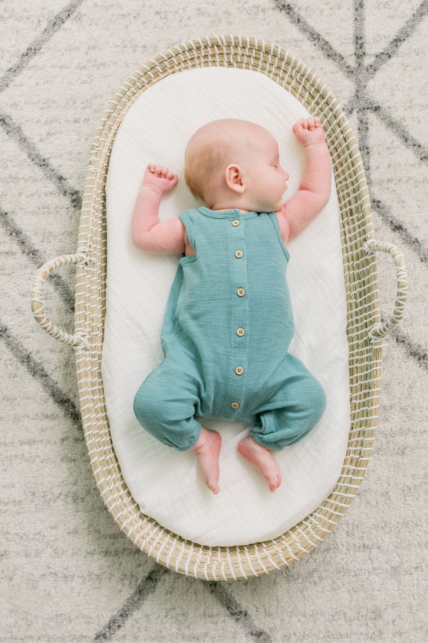 Baby wearing teal romper with buttons lying in woven bassinet on patterned rug.