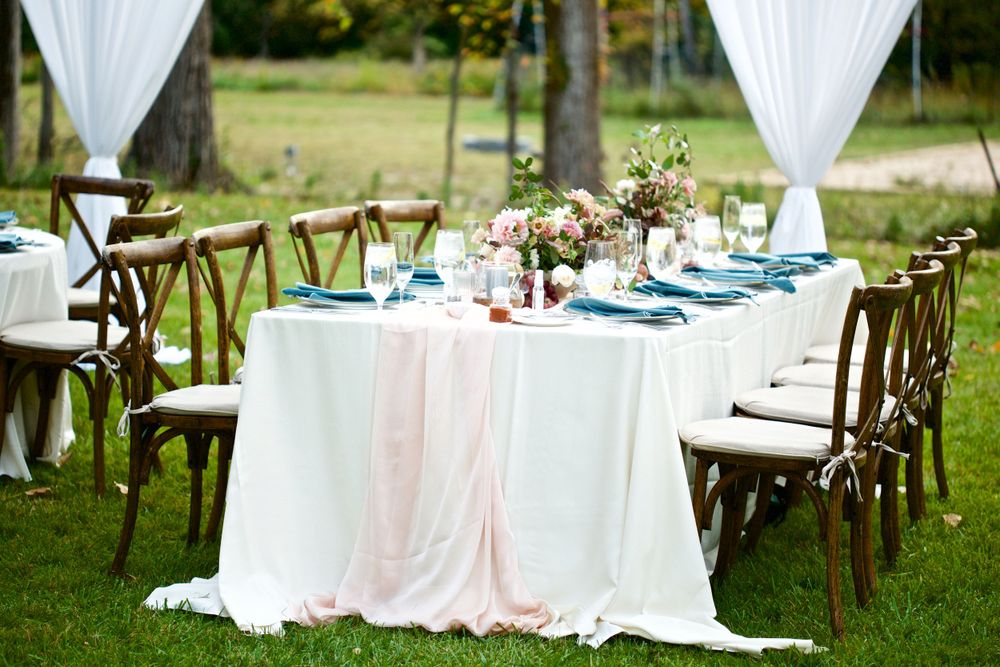 An outdoor wedding table setting with white linens, turquoise accents, and wooden cross-back chairs.