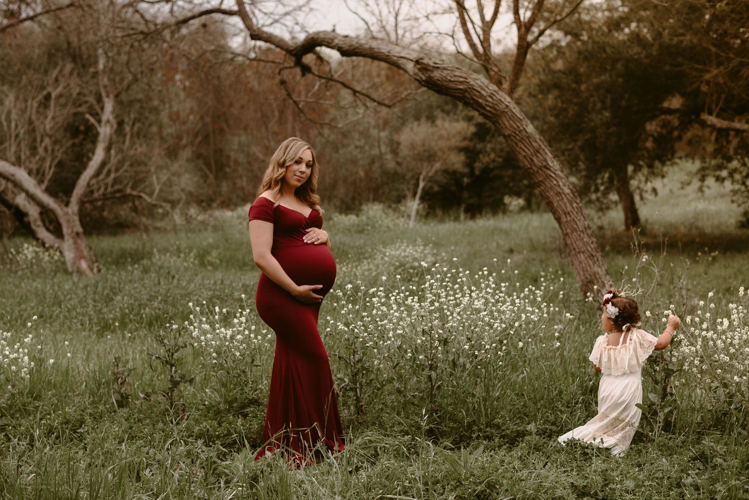 Maternity portrait in a field of wildflowers with a young child in white dress playing in the background.