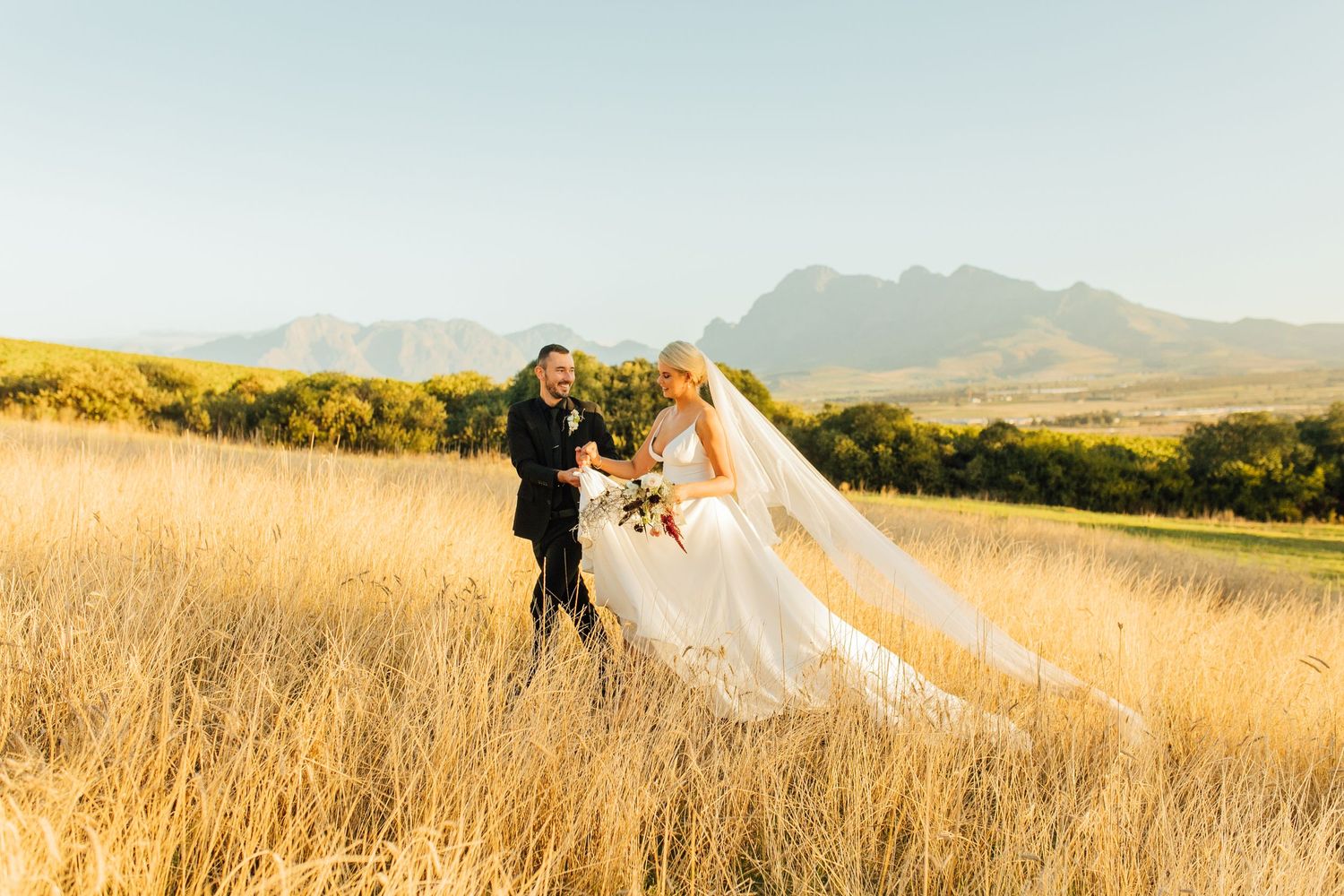 A newlywed couple stands together in a golden field with majestic mountains in the background at sunset.